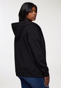 Kobieta o średnio-ciemnej skórze, ubrana w czarną bluzę z kapturem i niebieskie dżinsy, patrzy przez ramię na tle jasnego, jednolitego tła.