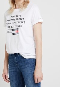 Hvid T-shirt med sort tekst, der står "EKT REAL KÆRLIGHED POSITIV ENERGI FORANDRING AF FREMTIDENÆGTE LYKKE" og et lille rektangulært logo nedenfor.