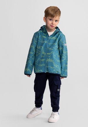 Jeune garçon portant une veste à capuche imprimée de dinosaures couleur sarcelle, un pantalon cargo marine et des baskets blanches, debout devant un fond gris clair.