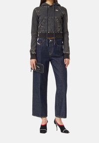 Žena má na sobě černou zkrácenou mikinu s kapucí, tmavě modré široké džíny s vysokým pasem, drží černou peněženku a nosí černé otevřené boty na Podpatku s červeným lemováním.