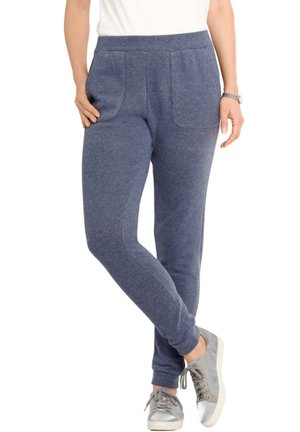 Frau, die blaue Jogginghosen mit elastischem Bund und Fronttaschen trägt, kombiniert mit silbernen Sneakers, steht mit einem Bein über das andere geschlagen.