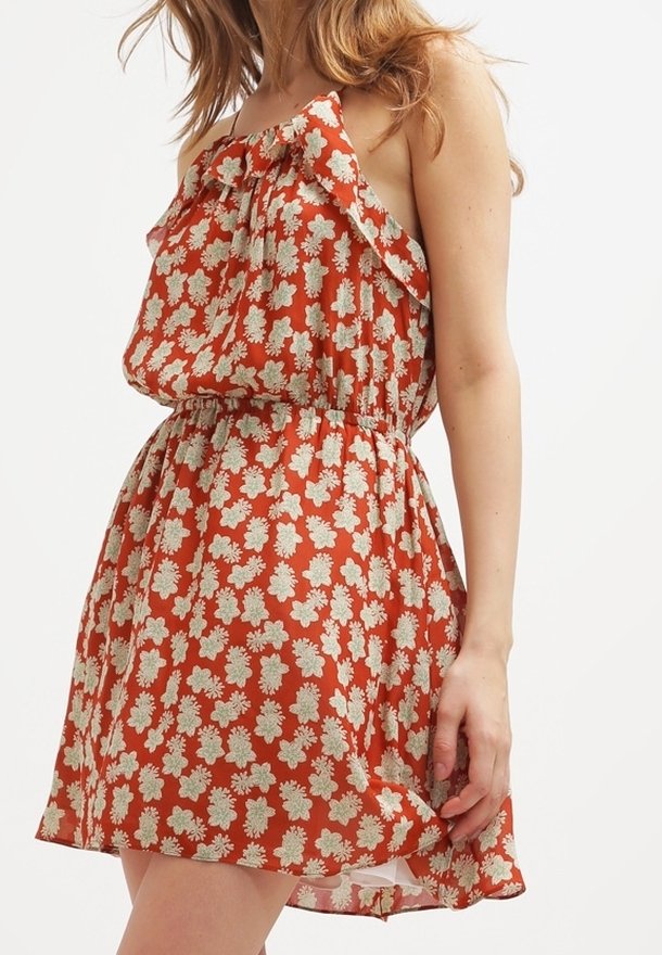 Femme portant une robe sans manches orange avec un motif floral blanc et un col à volants, debout devant un fond blanc.