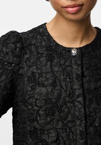 Svart teksturert jakke med et krøllete stoffdesign. Har rund hals og en blank knappelukking foran.