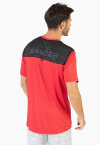 Camiseta deportiva roja con espalda de patrón de camuflaje negro y logo "SPYDER" en gris. Manga corta, textura suave, escote redondo, tela ligera.