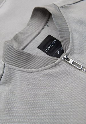 Sudadera gris con cremallera de tela texturizada, cuello acanalado y etiqueta de marca negra. Tiene una cremallera de metal y un diseño casual.