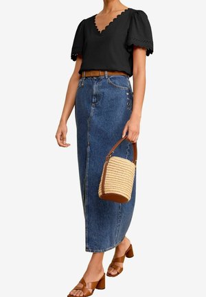 Vrouw draagt een zwart topje met gekartelde V-hals, een lange high-waist spijkerrok, een bruine riem, bruine sandalen met hak en houdt een geweven handtas vast.