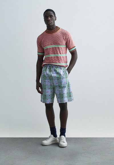 Κόκκινο πουλόβερ με υφή πλεξίματος με πράσινες και πορτοκαλί ρίγες, συνδυασμένο με καρό σορτς σε μπλε, πράσινο και άσπρο χρώμα· λευκά αθλητικά παπούτσια και ναυτικό μπλε κάλτσες.