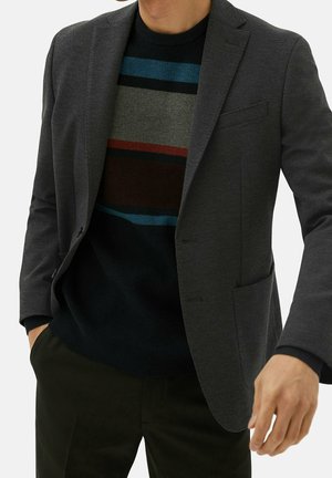 Homme portant un blazer gris foncé sur un pull rayé multicolore, les mains dans les poches, sur un fond uni.