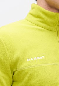 Φωτεινό κίτρινο φλις μπουφάν με ψηλό γιακά και κεντημένο λογότυπο "MAMMUT" στο στήθος, φορεμένο από άτομο που απεικονίζεται από το λαιμό έως τον ώμο.