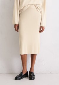 Jupe midi en tricot beige avec une taille ajustée. Associée à des mocassins en cuir noir, affichant une texture lisse et un design simple.