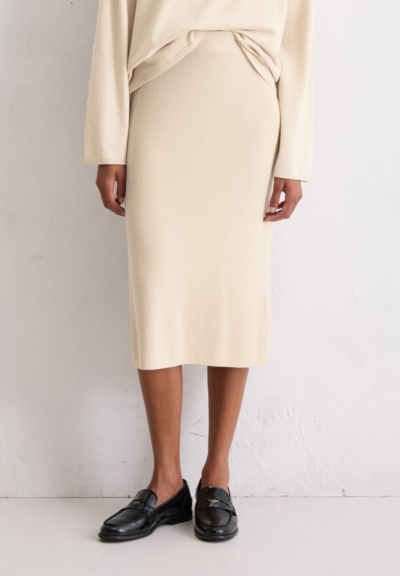 Jupe midi en tricot beige avec une taille ajustée. Associée à des mocassins en cuir noir, affichant une texture lisse et un design simple.