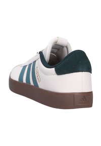 Zapatilla blanca con rayas en azul verde y talón de ante verde oscuro. Presenta un upper de cuero texturizado y una suela de goma marrón. Detalles en dorado en el lateral.