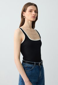 Top a maniche corte nero a coste con bordi scallop bianchi, caratterizzato da spalline larghe, scollatura quadrata e abbinato a jeans in denim blu a vita alta.