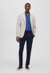 Blazer de lino beige claro con dos bolsillos delanteros sobre una camisa de mezclilla azul, combinado con pantalones navy y zapatillas oscuras marrones.