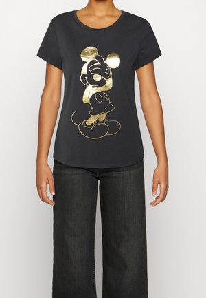 Osoba ubrana w czarną koszulkę z krótkim rękawem z metalicznym złotym nadrukiem Myszki Miki, w połączeniu z ciemnymi, szerokimi jeansami, na białym tle.