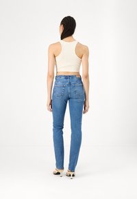 Femme aux cheveux tressés, de dos, portant un crop top blanc sans manches, un jean bleu taille haute et des talons beiges sur fond blanc.