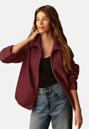 Jachetă fleece burgundy cu fermoar în față, guler mare și mâneci largi, purtată cu un top negru fără mâneci și blugi albaștri.