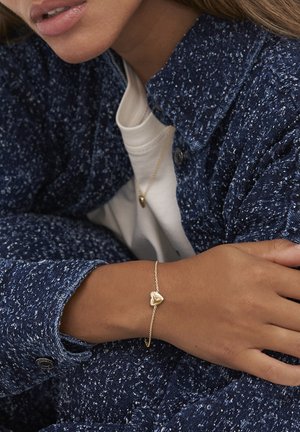 Primo piano di una persona che indossa un giubbotto navy blu a trama e una camicia bianca, con bracciale e collana a forma di cuore dorati coordinati.