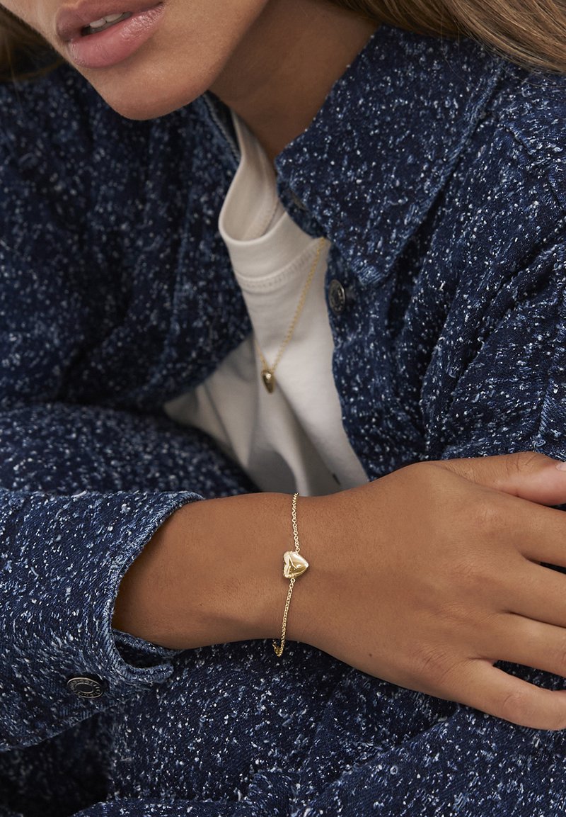 Primo piano di una persona che indossa un giubbotto navy blu a trama e una camicia bianca, con bracciale e collana a forma di cuore dorati coordinati.