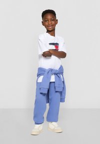 Weißes T-Shirt mit abstraktem roten und blauen Design, blaue Jogginghose und hellfarbige Sneakers. Sweatshirt um die Taille gebunden.