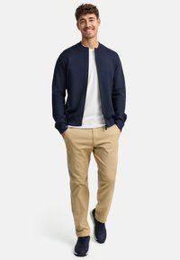 Chaqueta bomber de punto color navy, camiseta blanca, pantalones beiges y zapatillas navy. Modelo de pie con las manos en los bolsillos, fondo claro.