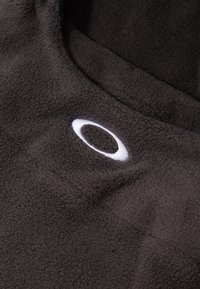 Hnedá fleece látka s výrazným bielym kruhovým logom. Mäkká textura s jemným leskom, ktorá odhaľuje hĺbku v záhyboch a vráskach.