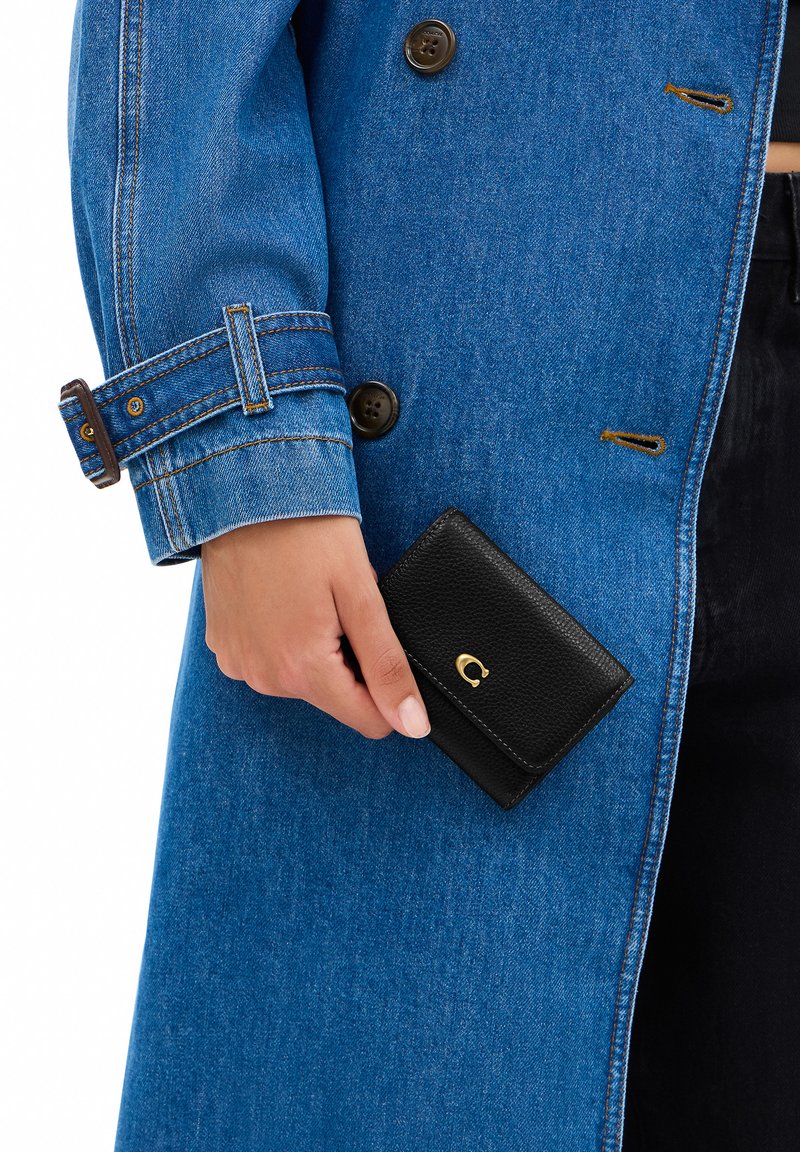 Main tenant un petit portefeuille en cuir noir avec fermeture dorée, portant un manteau en denim bleu avec des détails de boutons et de ceinture.