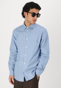 Camisa de botones de rayas azules con un solo bolsillo en el pecho, fabricada en tejido ligero; cuenta con un collar y mangas con puños. Gafas de sol oscuras puestas.