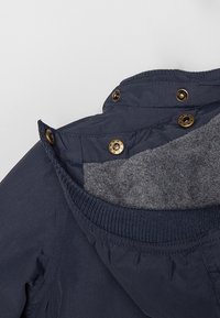 Gros plan sur le col d’une veste bleu marine montrant des boutons pression en laiton et une doublure en polaire grise à l’intérieur du col.