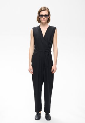 Femme portant une combinaison-pantalon noire sans manches portefeuille, des lunettes de soleil noires et des chaussures noires, debout devant un fond blanc uni.