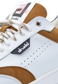 Zapatilla blanca con detalles de ante marrón. Presenta una parte superior texturizada, un diseño perforado y una distintiva etiqueta de logo negra. Cordoneras trenzadas.