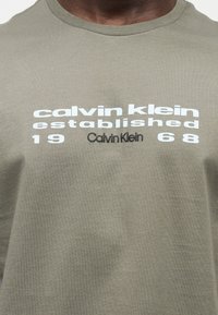 Κοντινό πλάνο ενός ατόμου που φορά γκρι-πράσινο πουκάμισο με λευκό και μαύρο λογότυπο "Calvin Klein established 1968" στο στήθος.