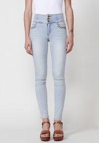 Jeans de cintura alta en azul claro con cierre de cinco botones, costuras decorativas en los bolsillos y un ajuste ceñido, combinados con sandalias de plataforma marrones.