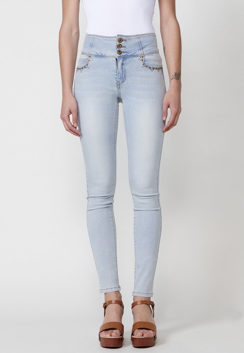 Jeans de cintura alta en azul claro con cierre de cinco botones, costuras decorativas en los bolsillos y un ajuste ceñido, combinados con sandalias de plataforma marrones.