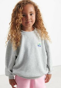 Heledus hall fliisist sviiter pika varrukaga ja ümmarguse kaelusega, millel on erksav värviline logoriba vasakul rinnal.