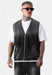 Schwarze Jeansweste mit V-Ausschnitt, Frontreißverschluss und zwei großen Taschen. Über einem weißen T-Shirt getragen, das einen abgeplatzten Look und rohe Kanten aufweist.