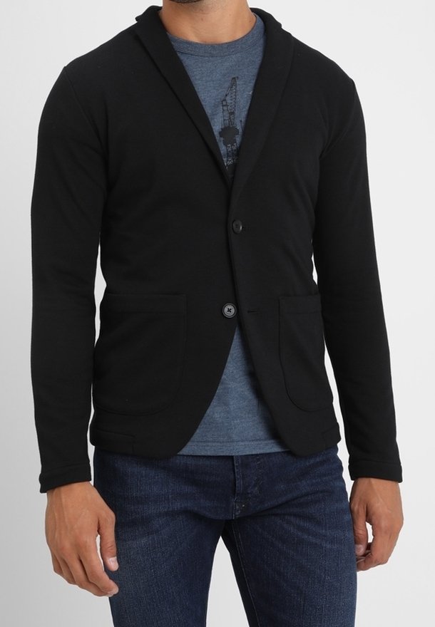 Homme portant un blazer noir boutonné sur un t-shirt bleu graphique et un jean bleu foncé, debout devant un fond uni.