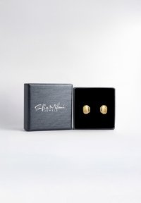 Guld øreringe i en sort smykkeboks med en blød indvendig flade. Boksen har en struktureret overflade og har et hvidt logo på forsiden.