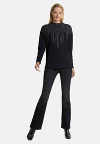 Zwarte top met lange mouwen, versierd met zilveren kraalaccenten in een verticaal patroon; gecombineerd met uitlopende zwarte jeans, versierd met verspreide zilveren accenten.