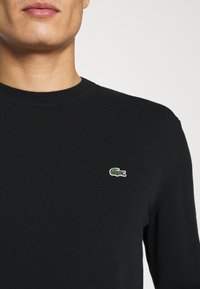 Suéter de punto negro con cuello redondo y textura acanalada, que presenta un pequeño logo de Lacoste en verde y blanco en el lado izquierdo del pecho.