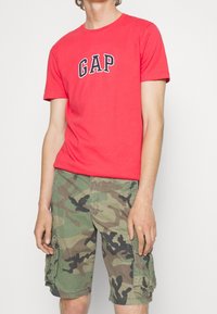 Persona vestida con una camiseta coral roja de GAP y pantalones cortos cargo verde camuflaje, de pie contra un fondo claro y liso.