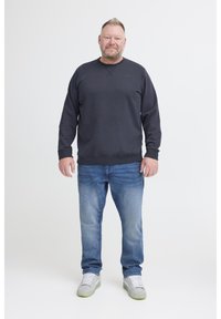 Férfi áll egy fehér hátt ér előtt, fekete kapucnis felsőt, kék farmert és világosszürke sportcipőt visel, fehér fűzőkkel.