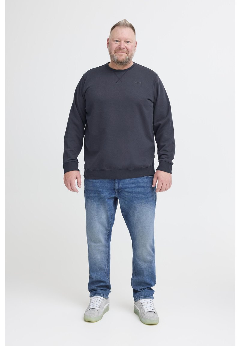 Férfi áll egy fehér háttér előtt, fekete kapucnis felsőt, kék farmert és világosszürke sportcipőt visel, fehér fűzőkkel.