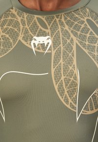 Olivgrünes, tailliertes Oberteil mit einem strukturierten Schlangenmuster in hellbeige. Verfügt über ein Emblehmotiv, das Zähnen ähnelt, im Brustbereich.