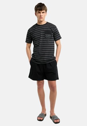 Jonge man staat met handen in zijn zakken, draagt een zwart gestreept bruno banani t-shirt, zwarte korte broek en grijze slippers op een witte achtergrond.