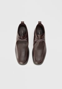 Zapatos de cuero marrón con un acabado texturizado, que presentan una punta redondeada, diseño con cordones y tirador en el talón. Suela plana.