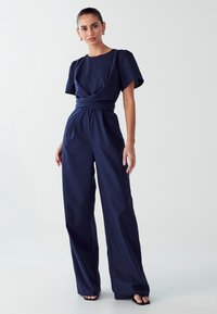 Mornarsko modra kombinezona s kratkimi napihnjenimi rokavi, ovitim pasom in širokimi hlačami. Narejena je iz teksturiranega materiala in ima gladko površino.