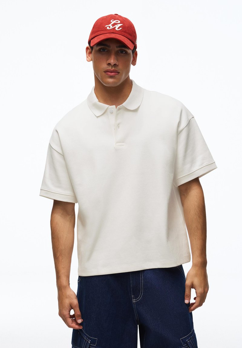 Jeune homme portant un polo blanc ample, un short en jean foncé et une casquette de baseball rouge avec des lettres blanches, debout devant un fond blanc uni.