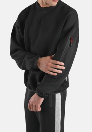 Mann trägt schwarzen Reebok-Sweatshirt mit Reißverschlusstasche am Ärmel und schwarze Jogginghose mit grauen Seitenpaneelen.