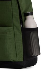 Mochila verde con bolsillo lateral de malla que contiene una botella blanca. El material es tela texturizada; cuenta con cremallera y detalles en negro.